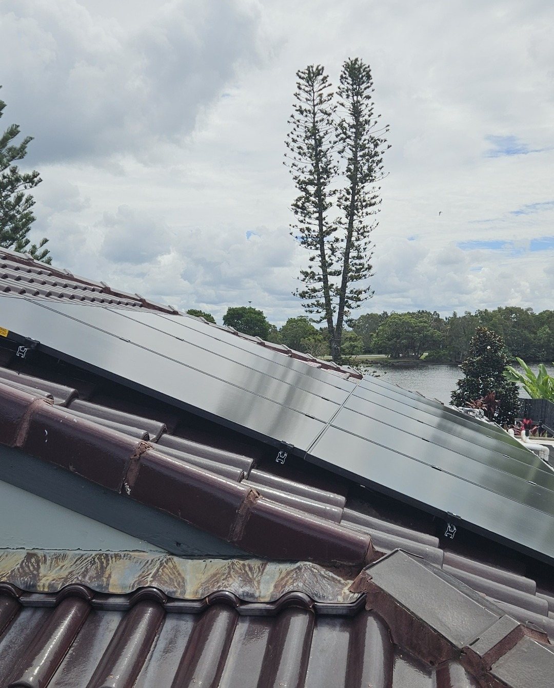 All-black AIKO solar panels on a terracotta tile roof in East Ballina All-black AIKO solar panels on a terracotta tile roof in East Ballina, with water and a tall pine behind, installed by Sun Connect Solar