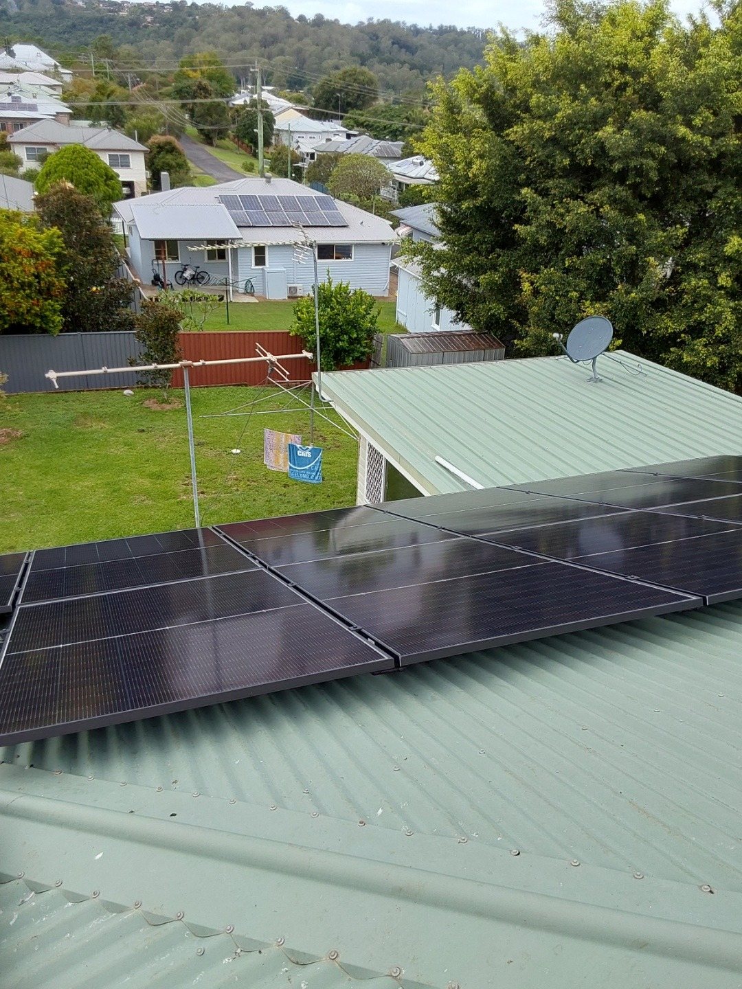 A row of Jinko solar panels on a green roof above a backyard in East Lismore, with houses and the hills behind, installed by Sun Connect Solar