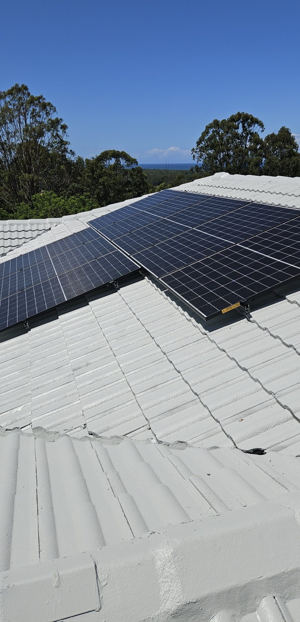 Jinko solar panels on a white tile roof in Ocean Shores with gum trees and a glimpse of the ocean behind Jinko solar panels on a white tile roof in Ocean Shores with gum trees and a glimpse of the ocean behind, part of a 16.92 kW install by Sun Connect Solar