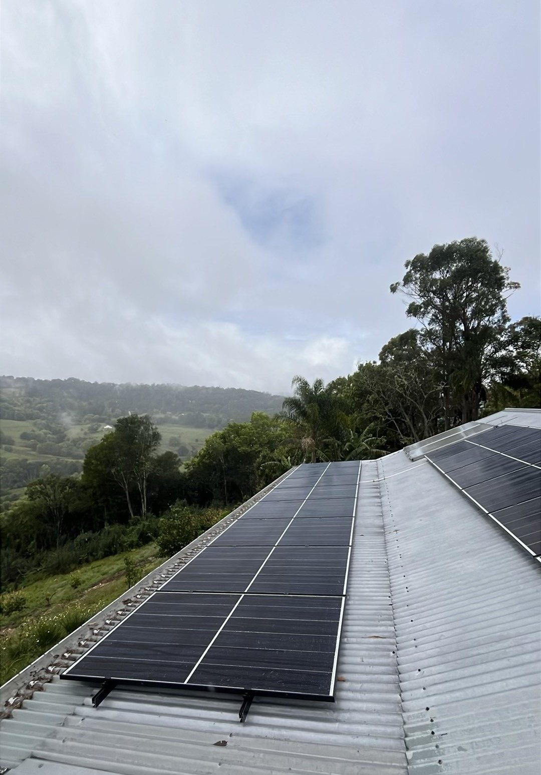 Jinko solar array on a corrugated roof with green hills behind at The Channon Jinko solar array on a corrugated roof with green hills behind at The Channon, part of a 6.65 kW solar and GoodWe battery install by Sun Connect Solar